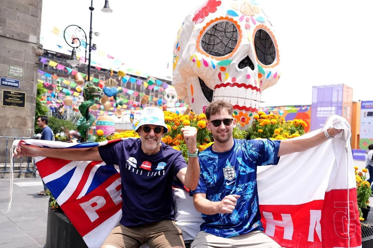 Two individuals, dressed in sports jerseys, are standing next to each other, holding flags and a decorative skull. They are seated in a festive street parade setting, with an audience in the background.